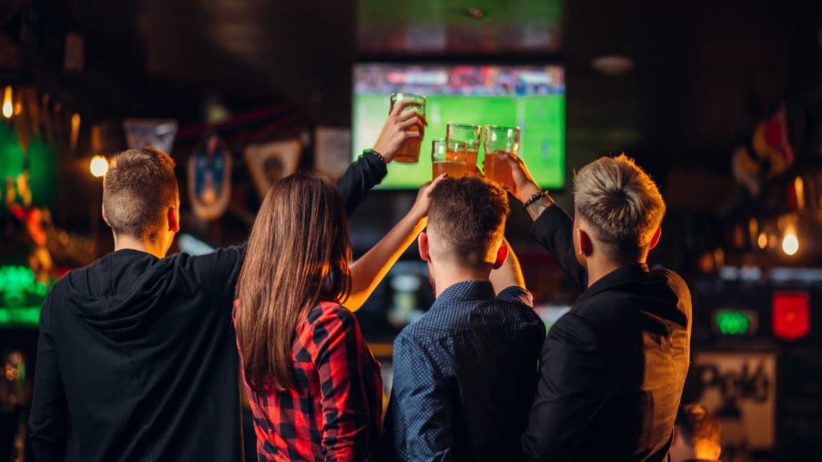A group of drinkers cheers their glasses in front of a screen showing the football in a bar